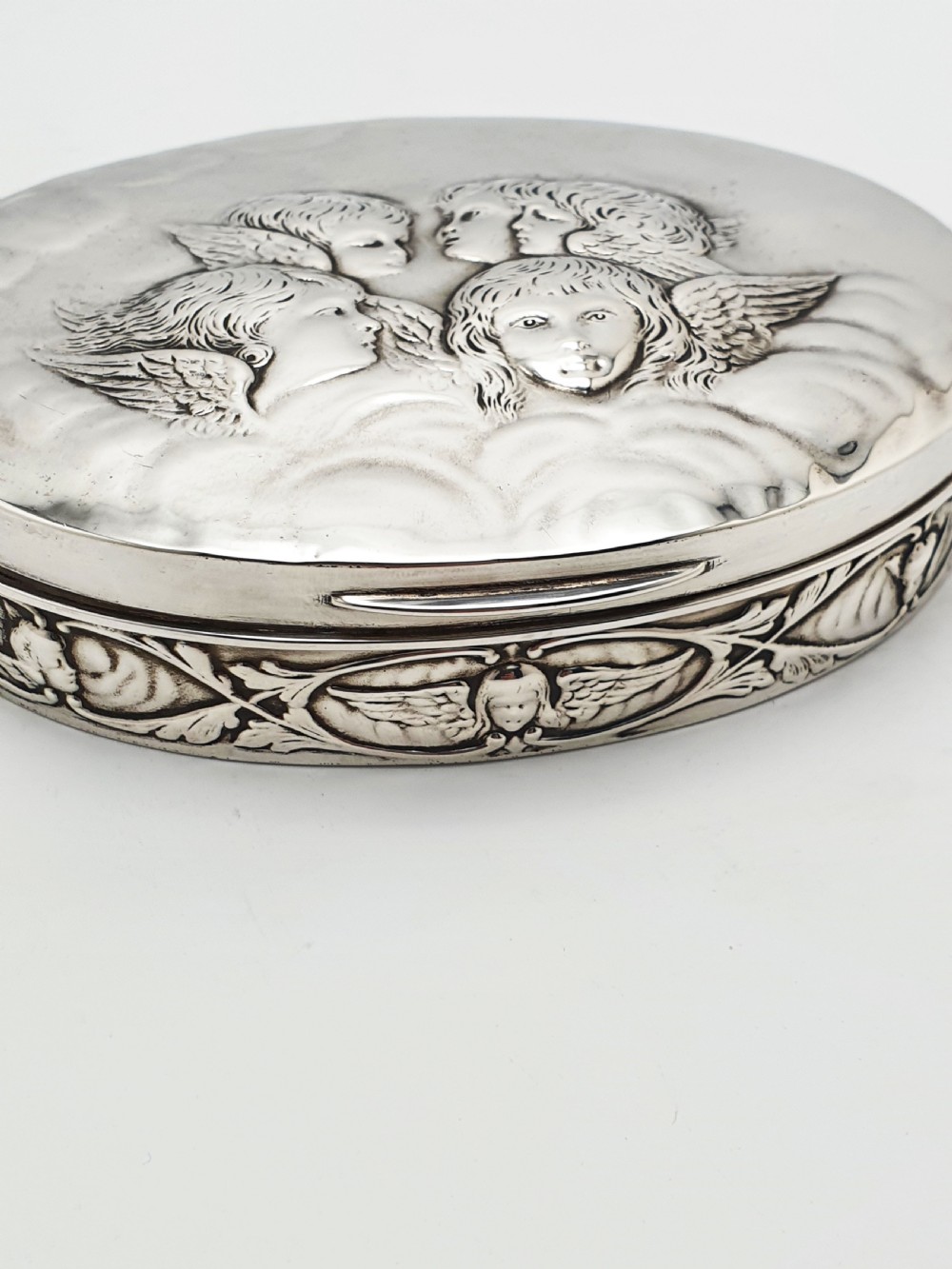 An oval sterling silver trinket box featuring an embossed lid with four cherub heads and a foliate border along the rim. View II