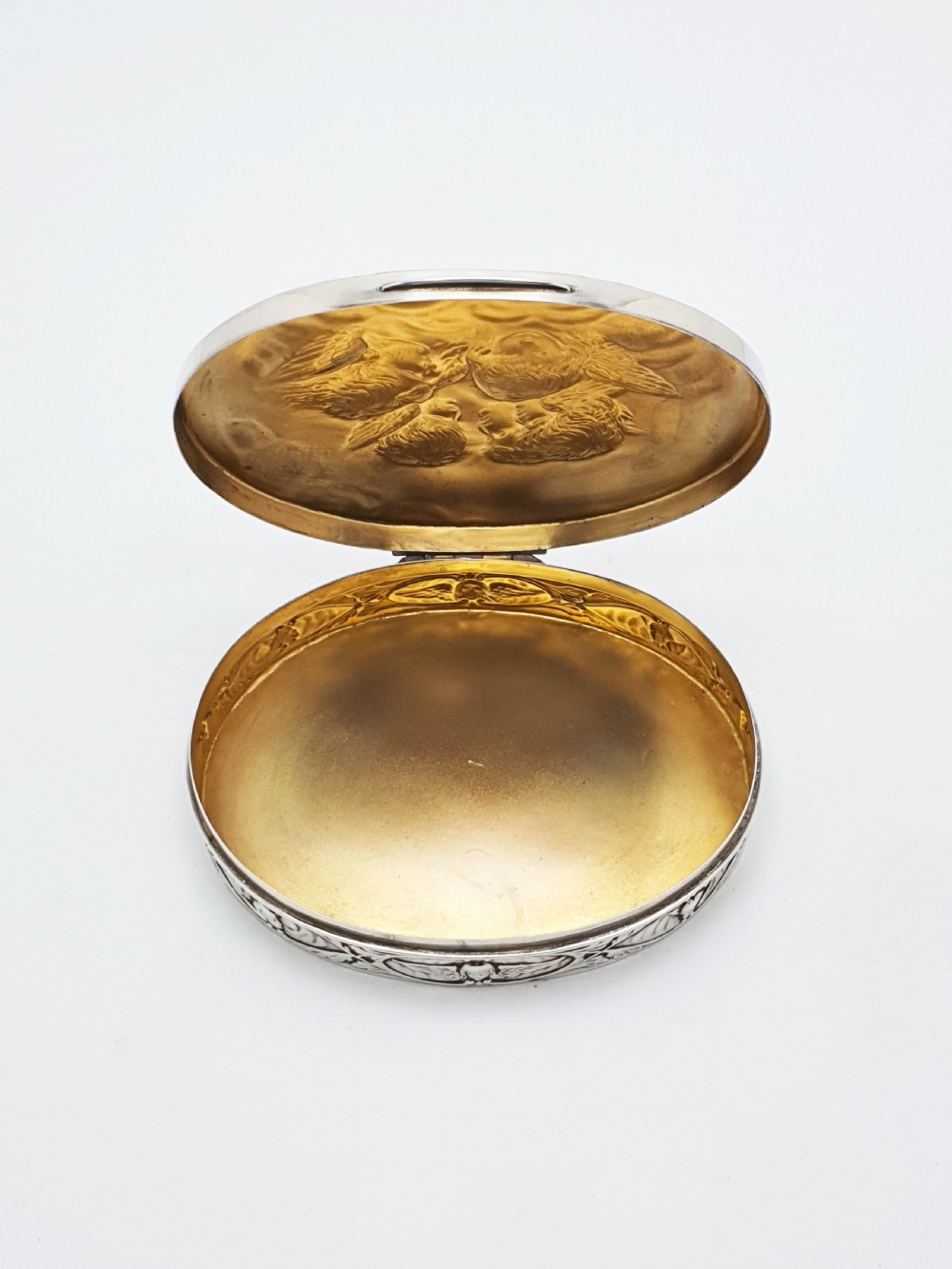 An oval sterling silver trinket box featuring an embossed lid with four cherub heads and a foliate border along the rim. View III