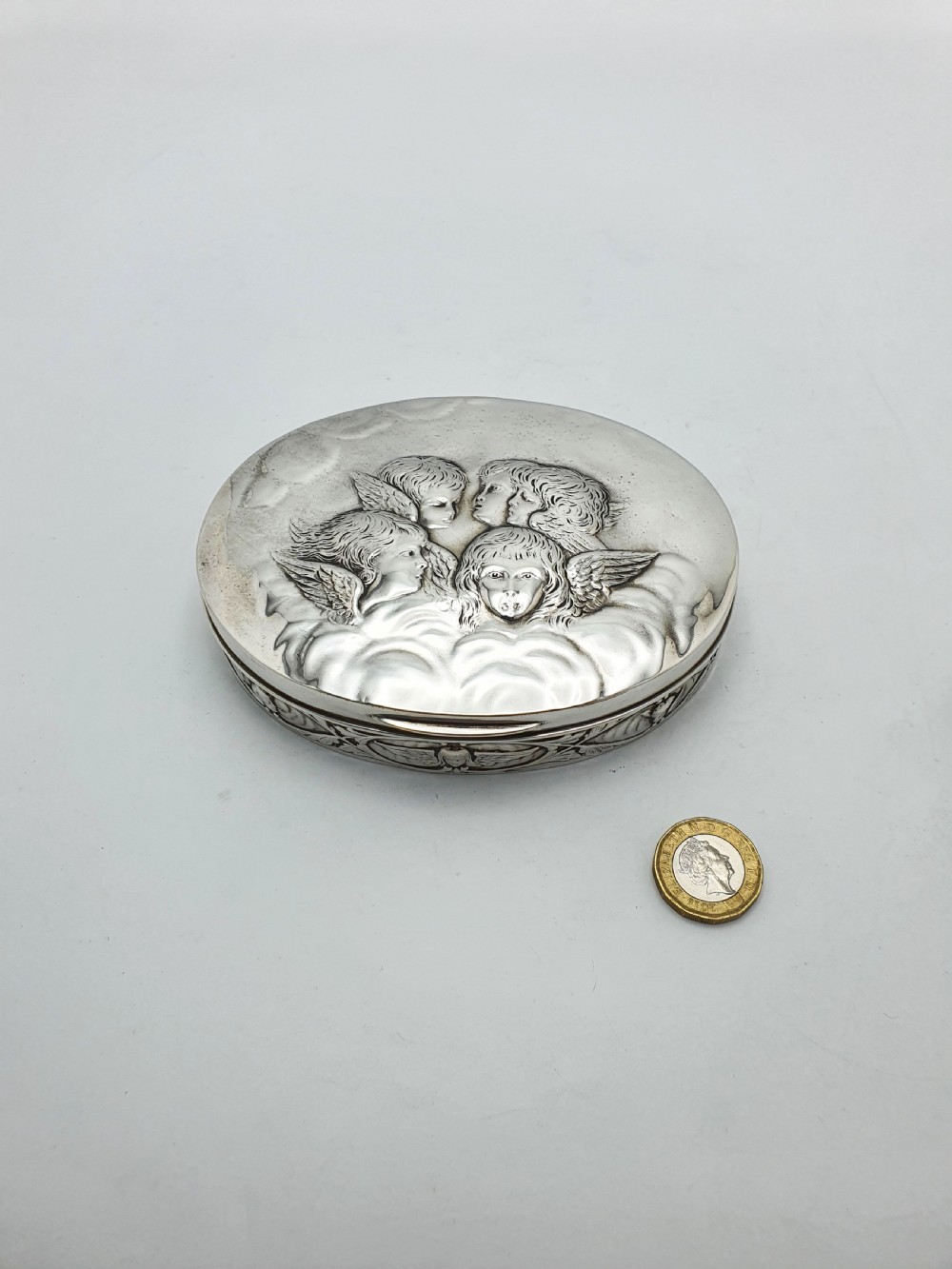 An oval sterling silver trinket box featuring an embossed lid with four cherub heads and a foliate border along the rim. View VII