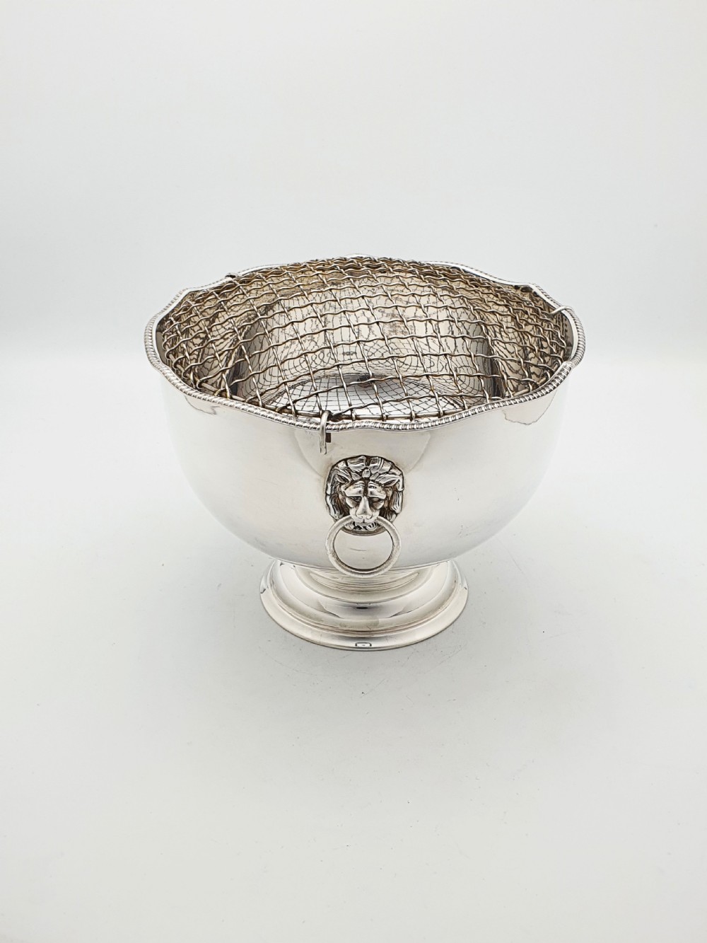 A circular silver-plated rose bowl featuring lion mask ring handles, a stepped pedestal base, and a removable wire mesh grid insert.