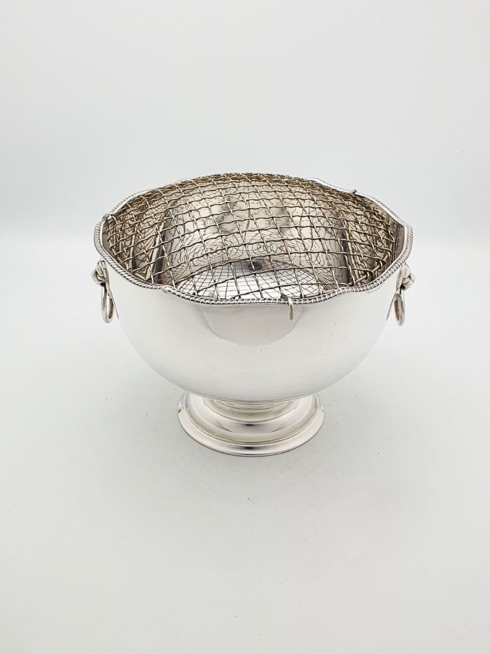 A circular silver-plated rose bowl featuring lion mask ring handles, a stepped pedestal base, and a removable wire mesh grid insert. View II