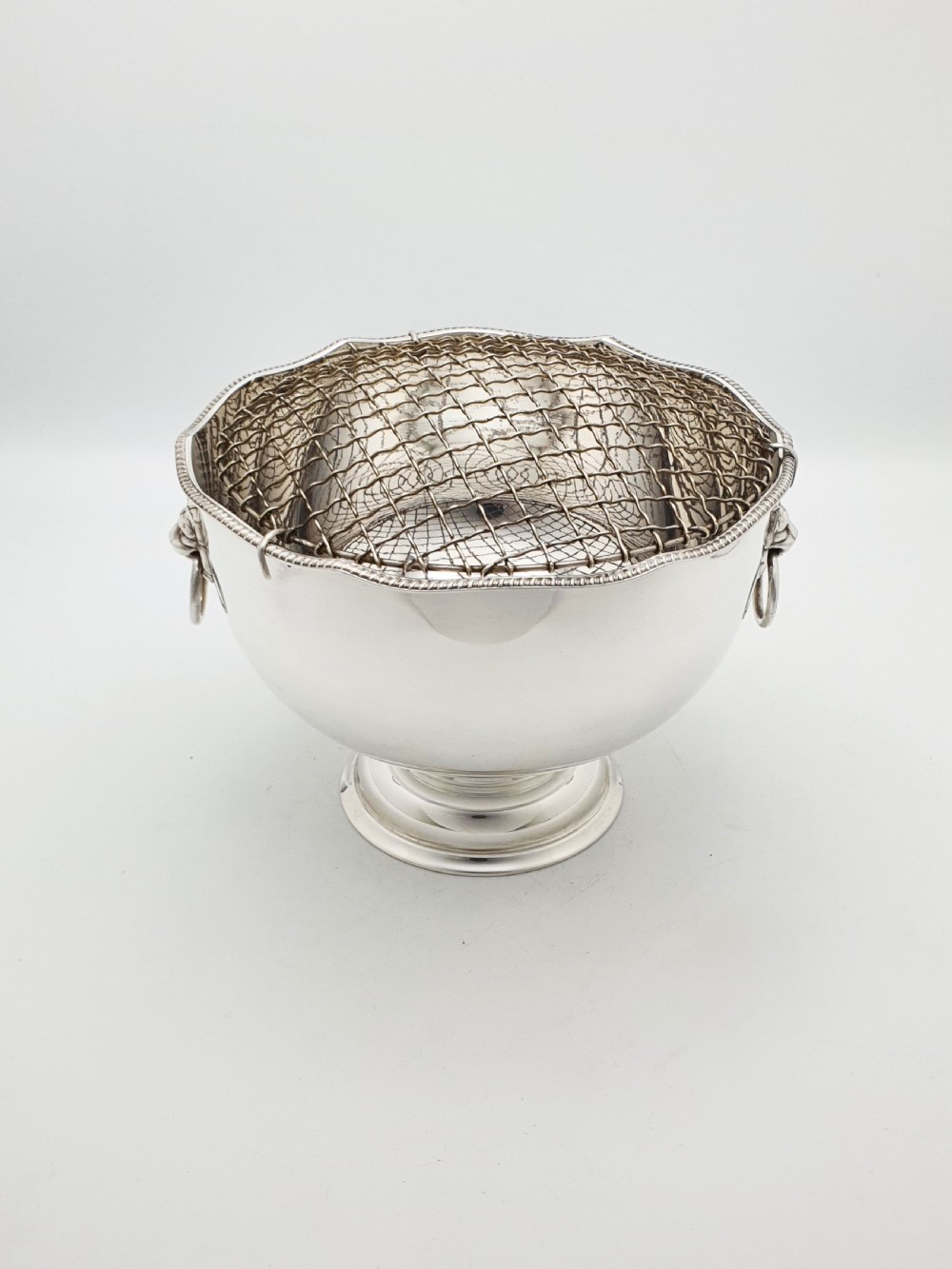 A circular silver-plated rose bowl featuring lion mask ring handles, a stepped pedestal base, and a removable wire mesh grid insert. View V