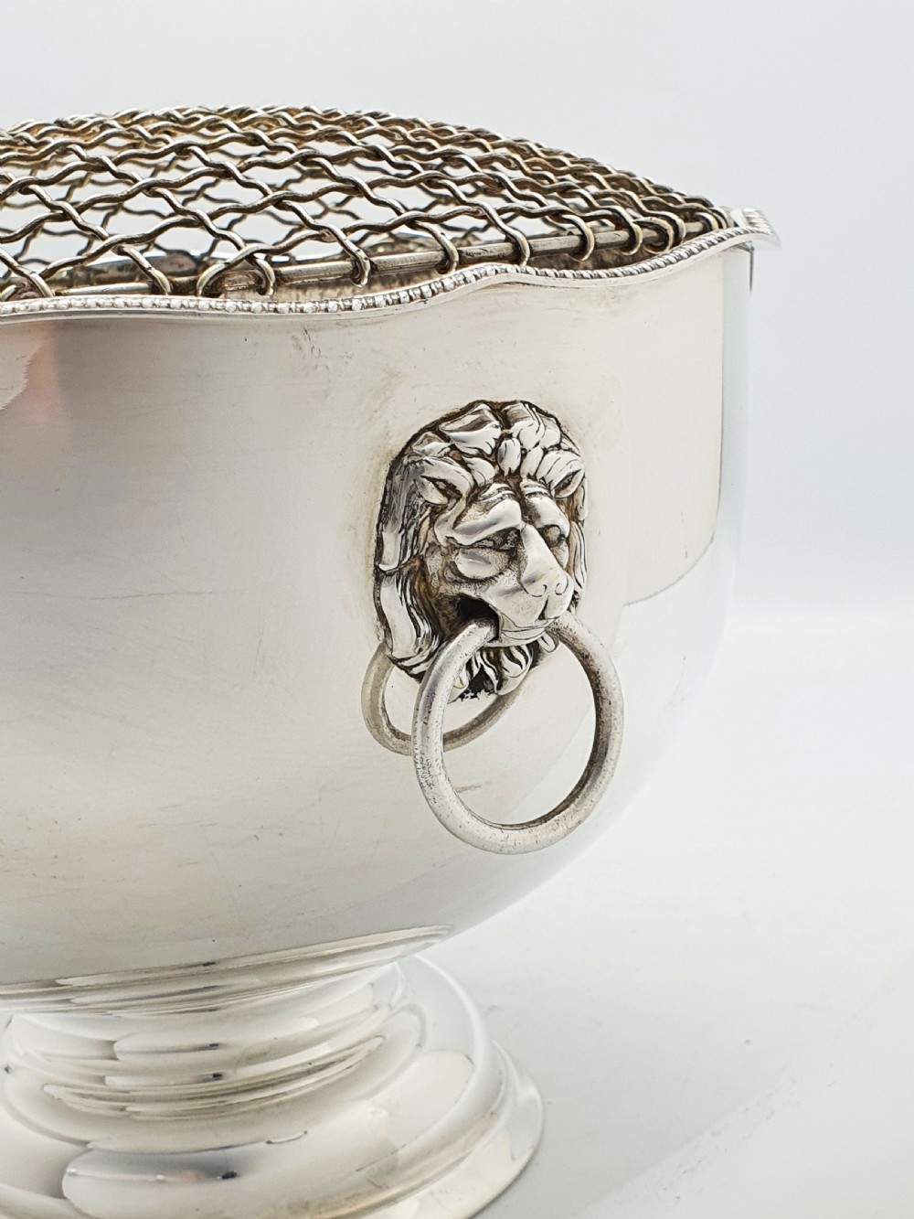 A circular silver-plated rose bowl featuring lion mask ring handles, a stepped pedestal base, and a removable wire mesh grid insert. View VI