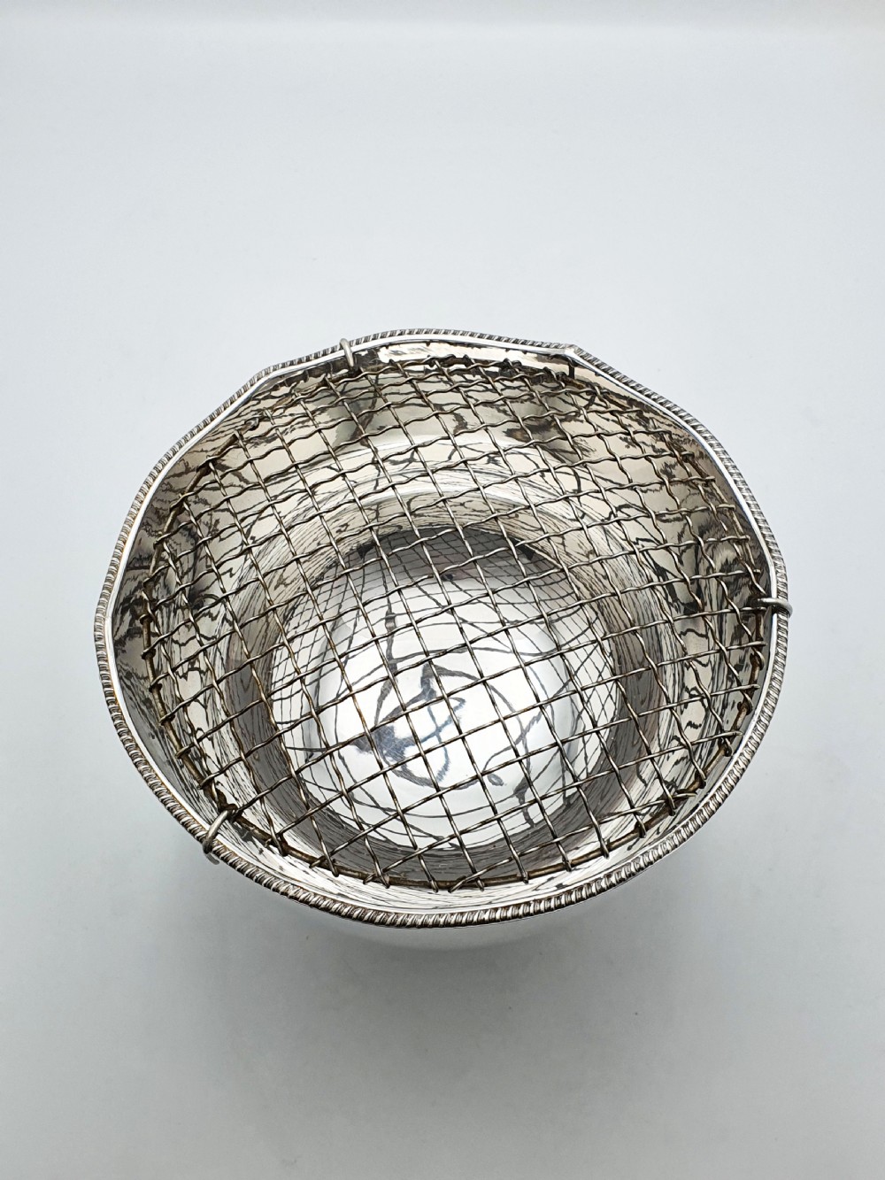 A circular silver-plated rose bowl featuring lion mask ring handles, a stepped pedestal base, and a removable wire mesh grid insert. View VII