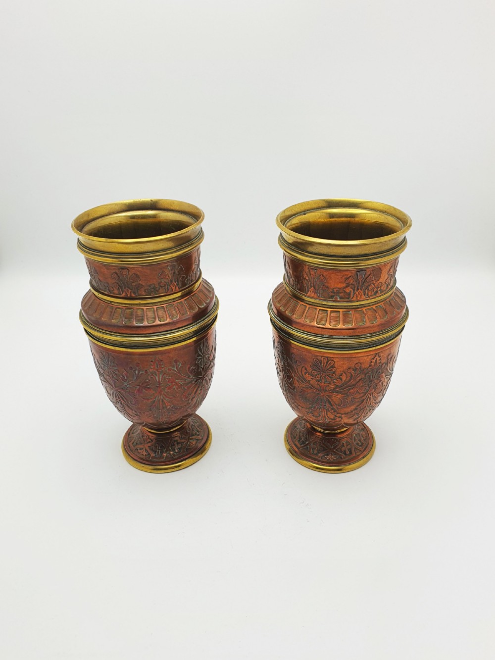 A pair of antique French copper urns featuring brass rims, pedestal bases, and chased floral motifs.