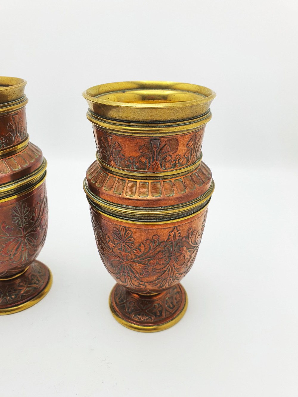 A pair of antique French copper urns featuring brass rims, pedestal bases, and chased floral motifs. View V