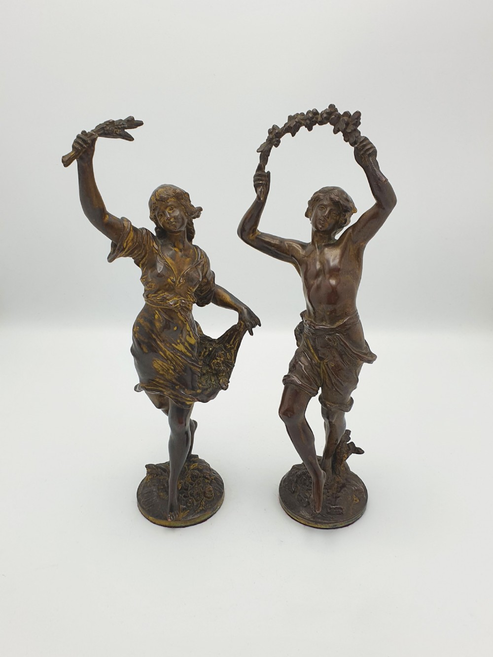 A pair of dark patinated bronze figurines depicting male and female harvest dancers holding floral garlands.