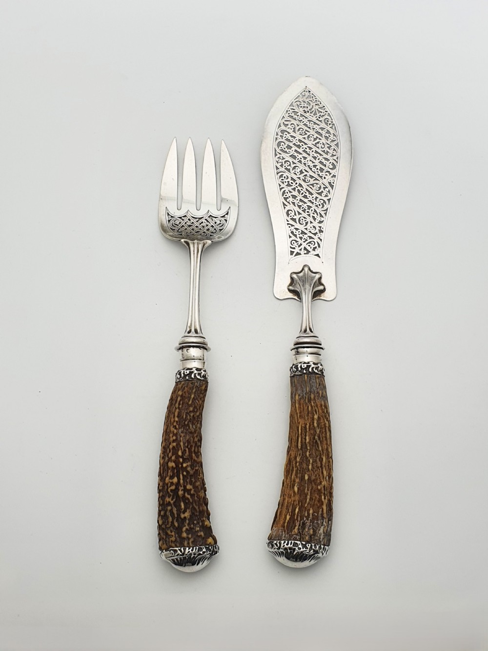 A pair of Victorian silver fish servers featuring intricately engraved pierced blades and natural stag antler handles.