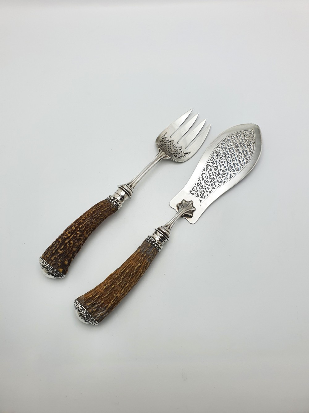 A pair of Victorian silver fish servers featuring intricately engraved pierced blades and natural stag antler handles. View II