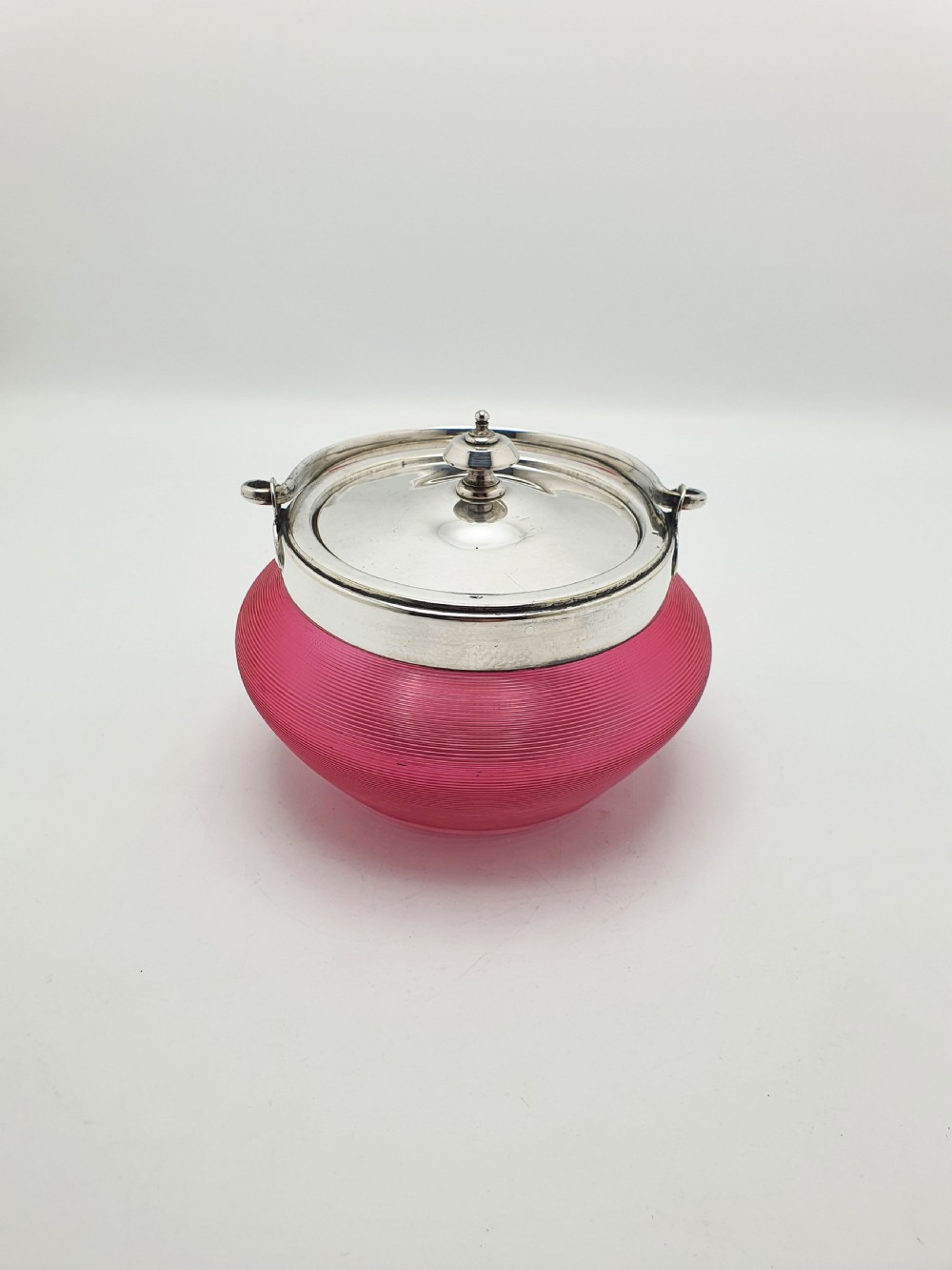 A ribbed cranberry glass biscuit barrel featuring a silver-plated fitted rim, flat lid, and hinged swing handle.