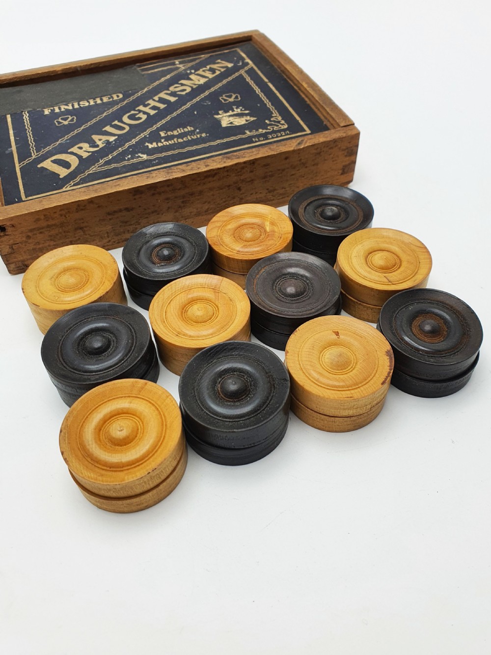 A set of turned boxwood and dark-stained wooden draughtsmen displayed in front of their original wooden slide-lid box with a printed label.