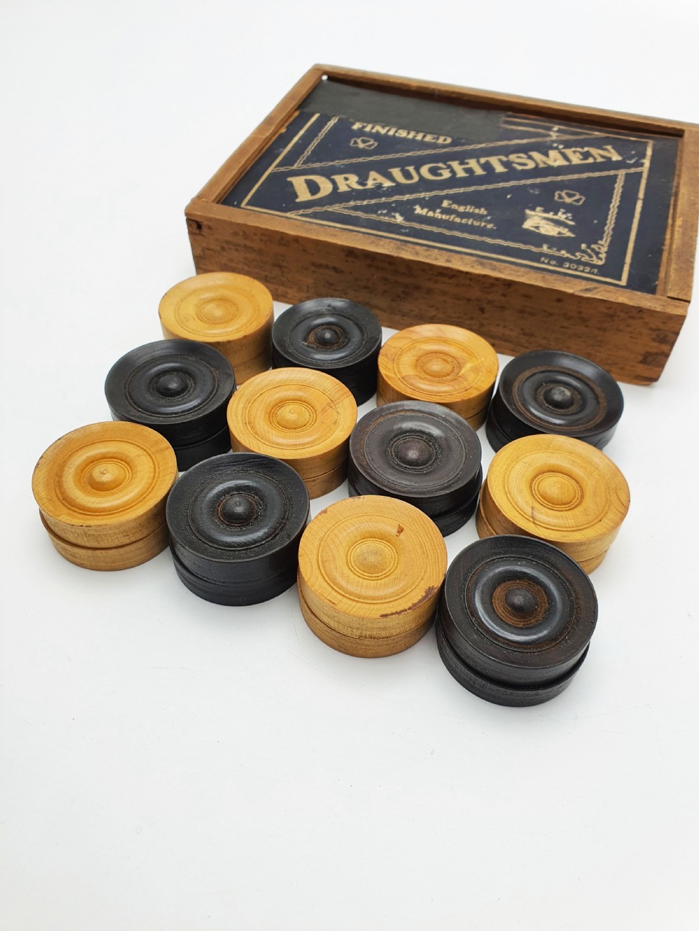 A set of turned boxwood and dark-stained wooden draughtsmen displayed in front of their original wooden slide-lid box with a printed label. View III