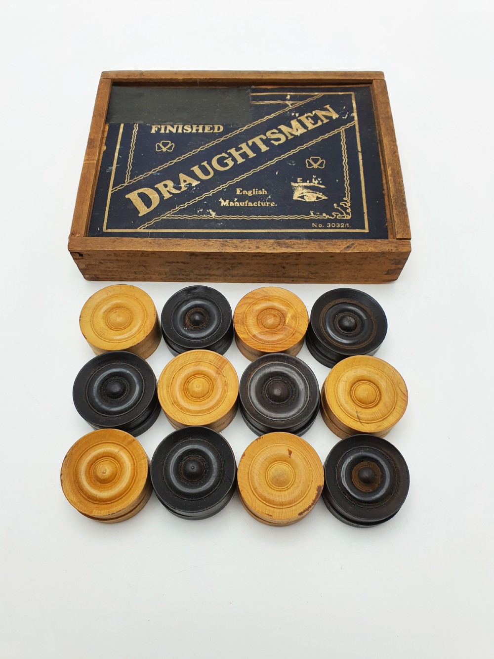 A set of turned boxwood and dark-stained wooden draughtsmen displayed in front of their original wooden slide-lid box with a printed label. View VI