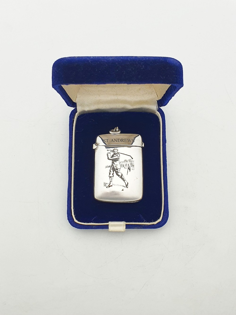 An antique sterling silver vesta case engraved with a golfer and St. Andrews lettering, displayed inside an open blue velvet box.
