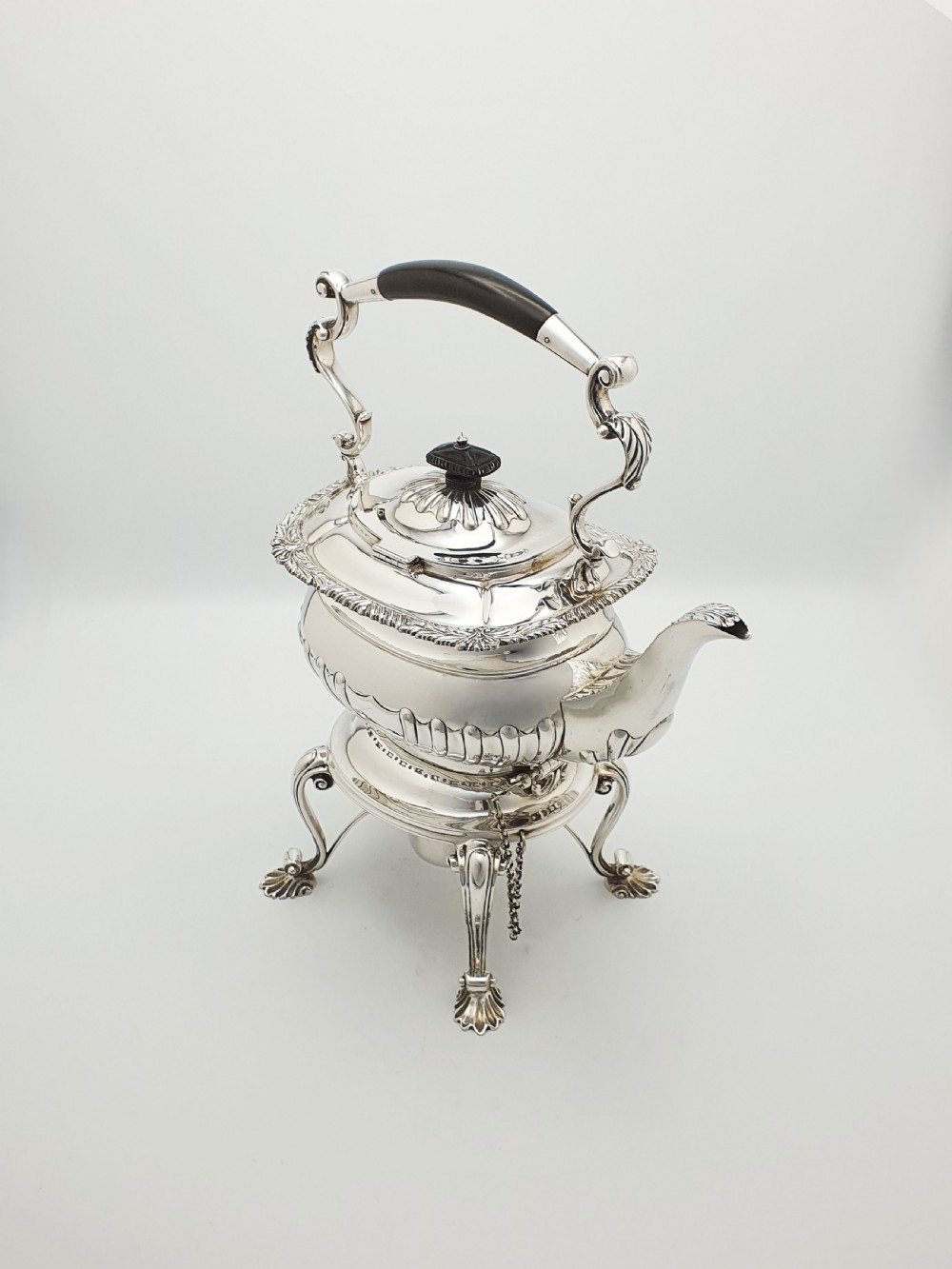 An antique Georgian-style silver-plated spirit kettle resting on a three-legged burner stand with shell feet and an ebony handle.