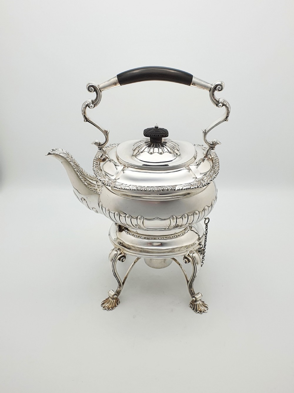 An antique Georgian-style silver-plated spirit kettle resting on a three-legged burner stand with shell feet and an ebony handle. View II