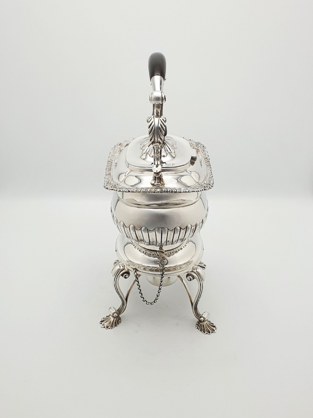 An antique Georgian-style silver-plated spirit kettle resting on a three-legged burner stand with shell feet and an ebony handle. View III