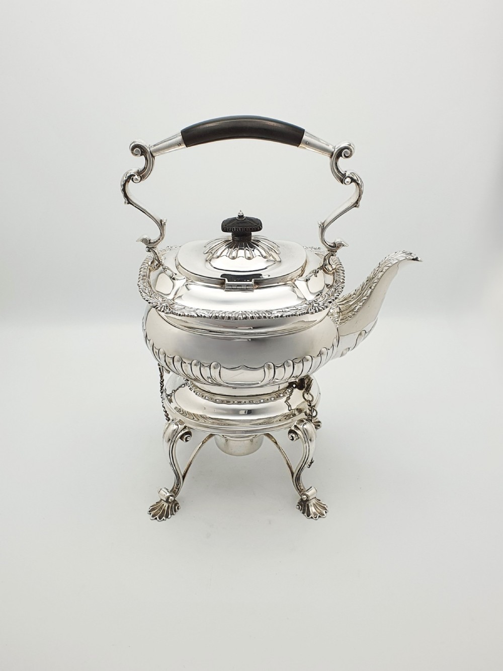 An antique Georgian-style silver-plated spirit kettle resting on a three-legged burner stand with shell feet and an ebony handle. View V