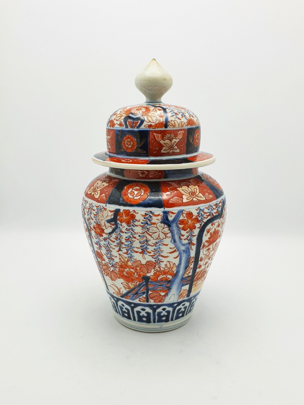 A Japanese Imari porcelain covered ginger jar decorated with hand-painted iron-red and cobalt-blue floral panels, complete with a domed lid and pointed finial.