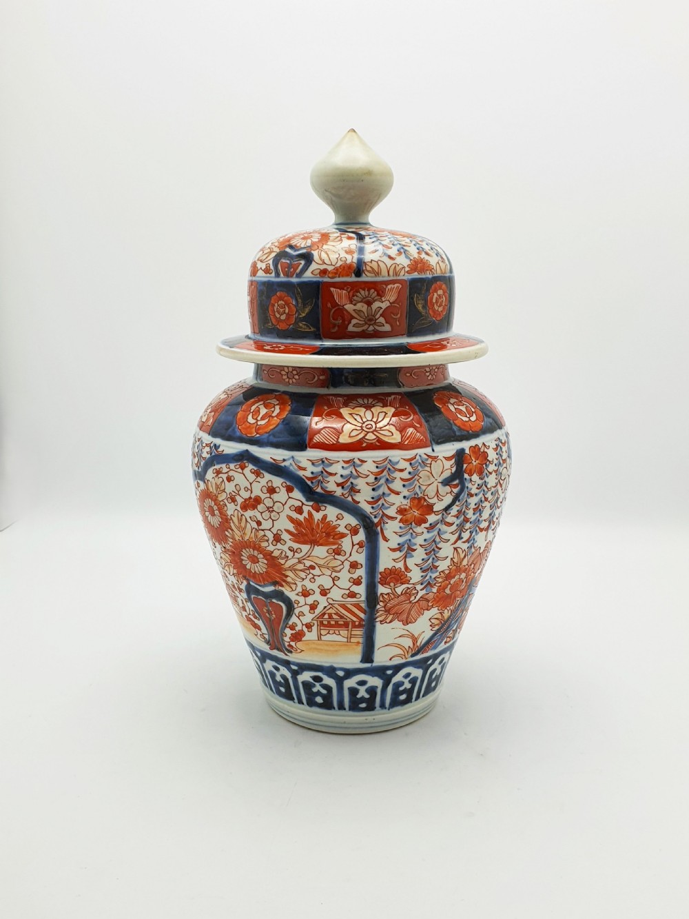 A Japanese Imari porcelain covered ginger jar decorated with hand-painted iron-red and cobalt-blue floral panels, complete with a domed lid and pointed finial. View IX