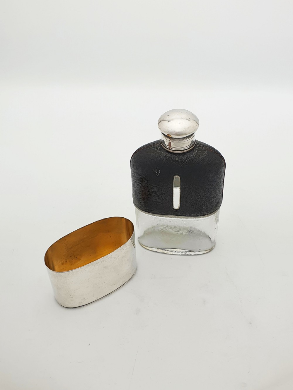 An Edwardian hip flask featuring a dark brown textured leather sleeve with a sight window, a removable silver-plated base cup, and a rounded silver-plated cap. View VI