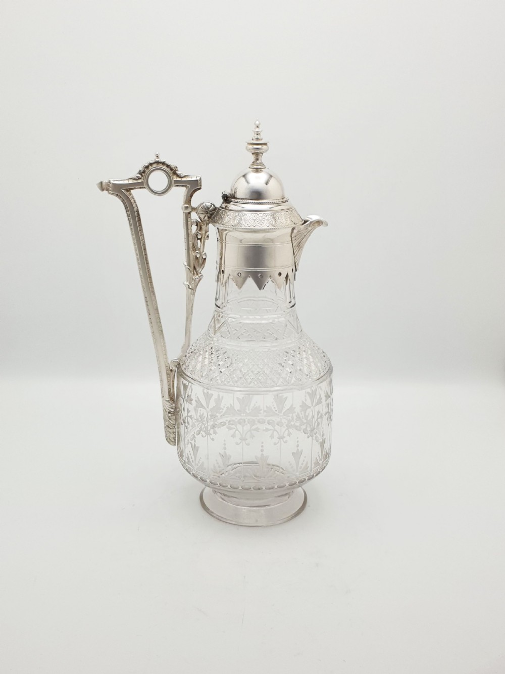 A late Victorian silver-plated and clear cut-glass claret jug with an engraved foliate body, domed hinged lid, and decorative angular handle.