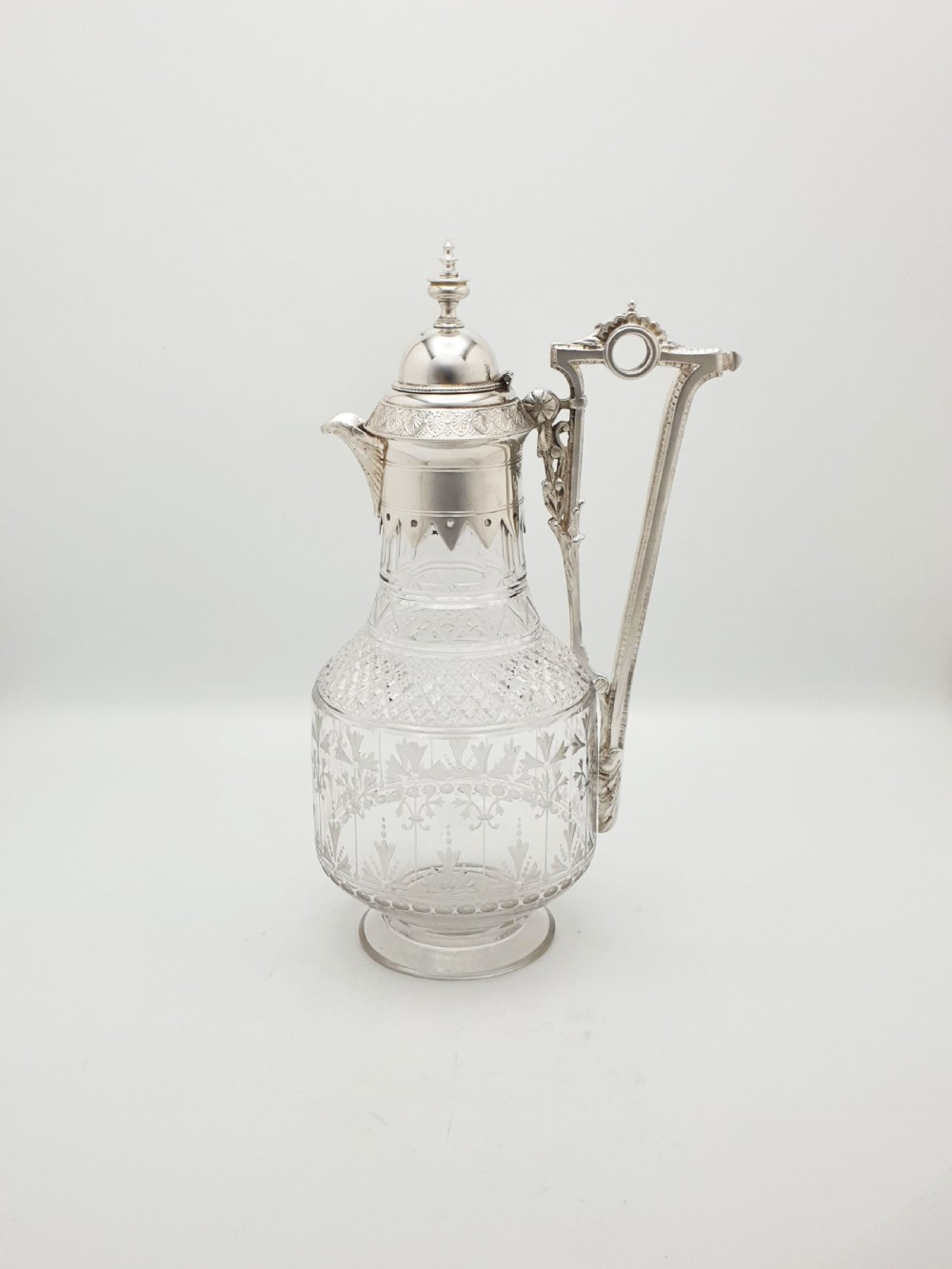 A late Victorian silver-plated and clear cut-glass claret jug with an engraved foliate body, domed hinged lid, and decorative angular handle. View II