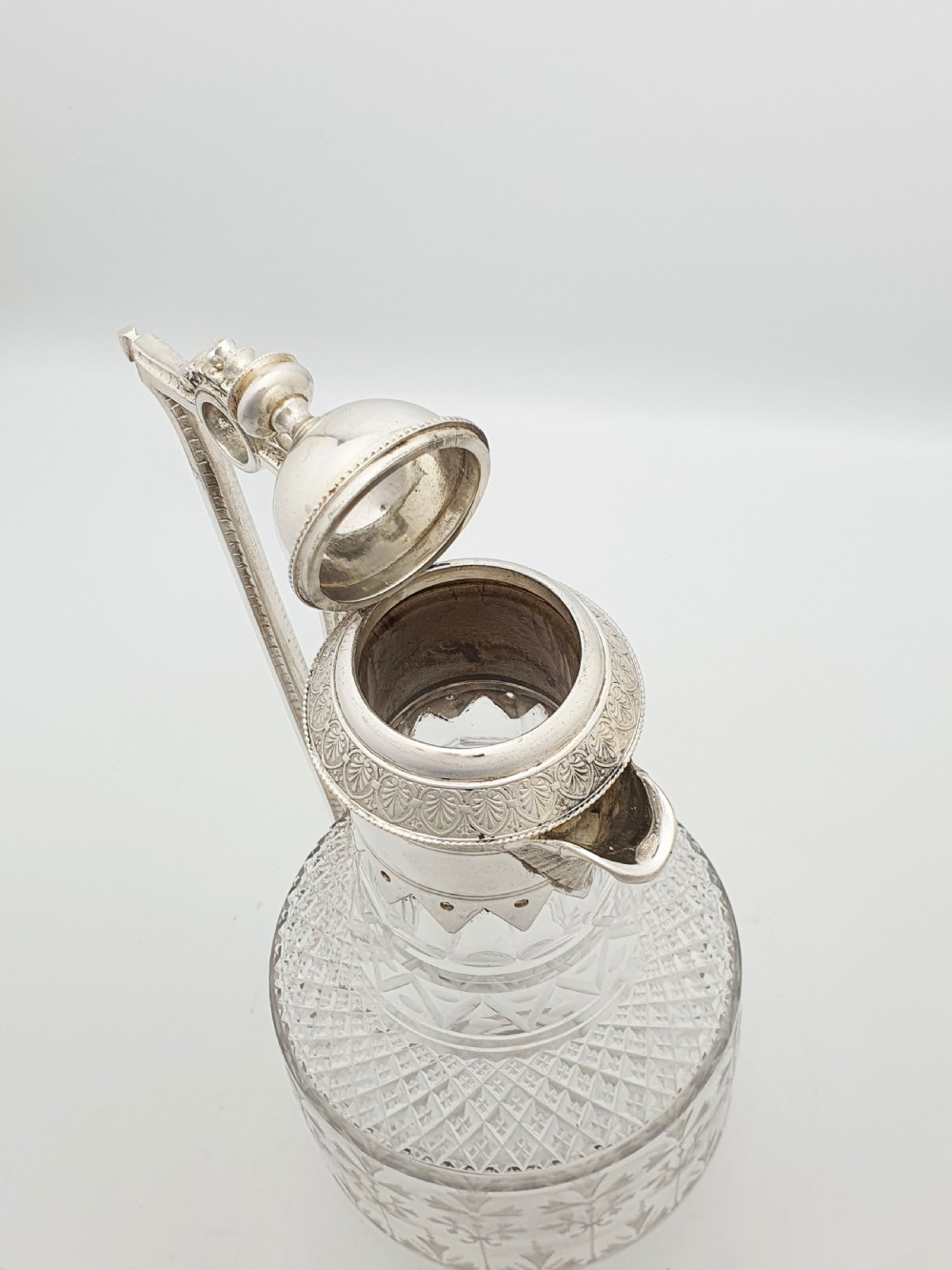 A late Victorian silver-plated and clear cut-glass claret jug with an engraved foliate body, domed hinged lid, and decorative angular handle. View V