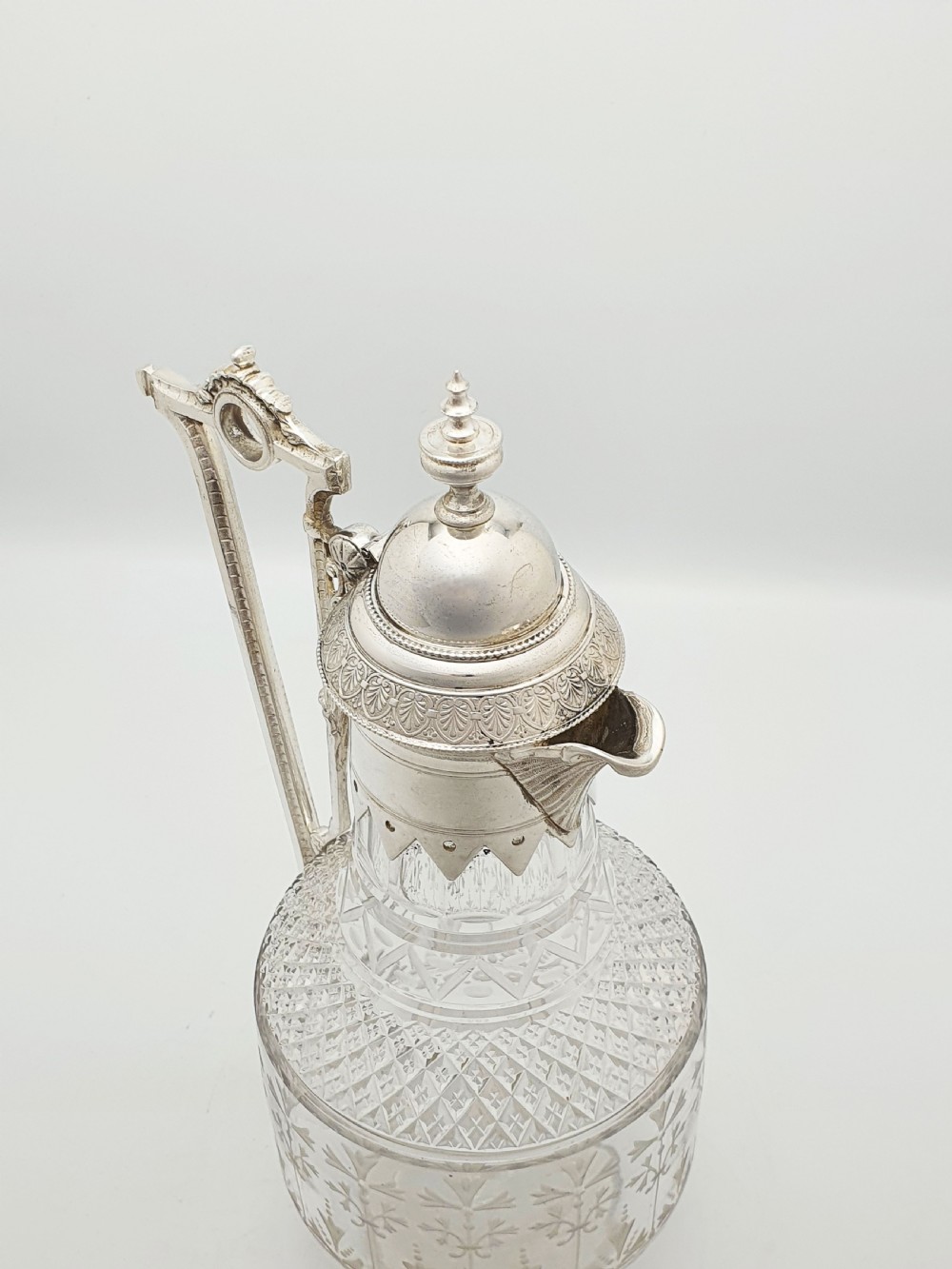 A late Victorian silver-plated and clear cut-glass claret jug with an engraved foliate body, domed hinged lid, and decorative angular handle. View VI