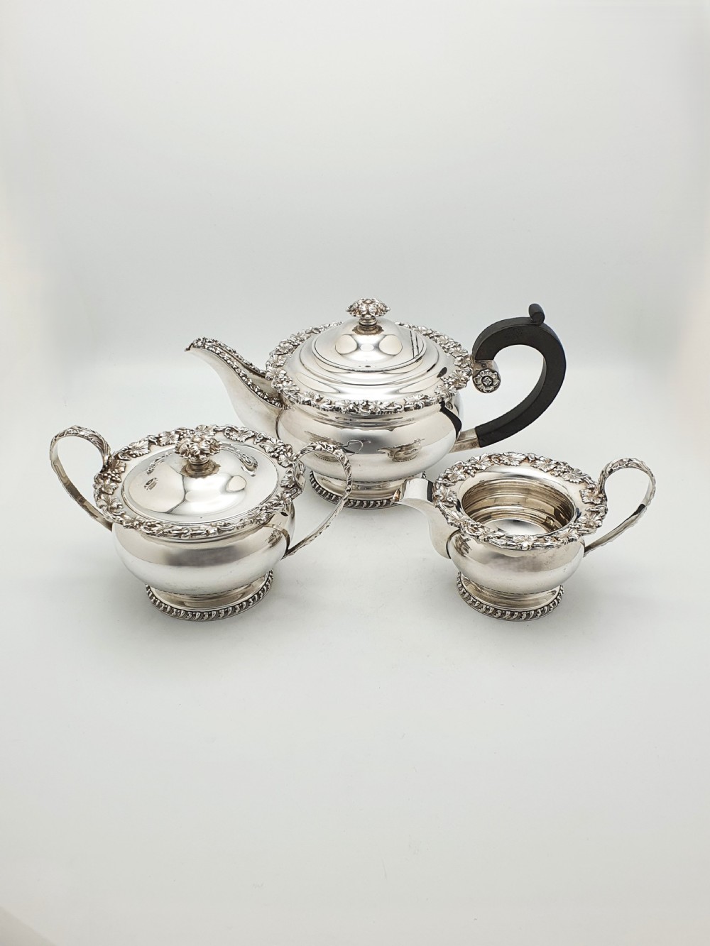 A three-piece silver-plated tea set featuring a teapot with a dark wood handle, a matching sugar bowl with lid, and a cream jug, all decorated with raised grapevine borders.