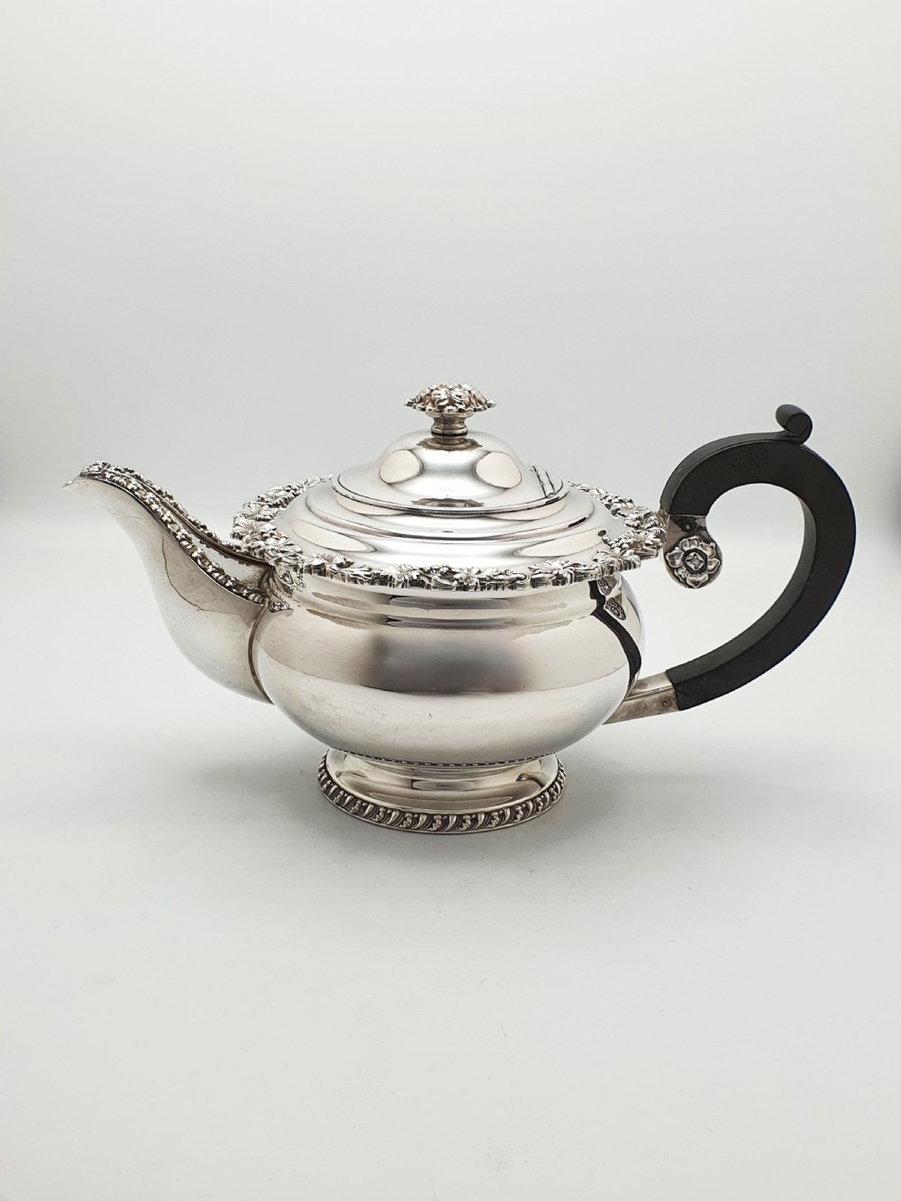 A three-piece silver-plated tea set featuring a teapot with a dark wood handle, a matching sugar bowl with lid, and a cream jug, all decorated with raised grapevine borders. View II