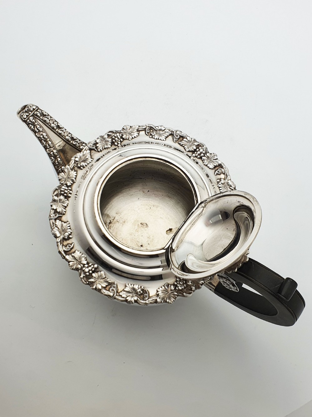 A three-piece silver-plated tea set featuring a teapot with a dark wood handle, a matching sugar bowl with lid, and a cream jug, all decorated with raised grapevine borders. View III