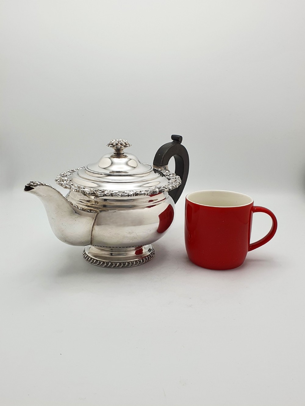 A three-piece silver-plated tea set featuring a teapot with a dark wood handle, a matching sugar bowl with lid, and a cream jug, all decorated with raised grapevine borders. View V