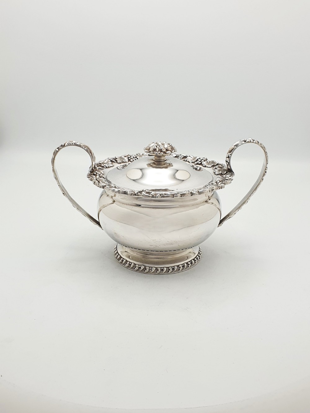 A three-piece silver-plated tea set featuring a teapot with a dark wood handle, a matching sugar bowl with lid, and a cream jug, all decorated with raised grapevine borders. View VI