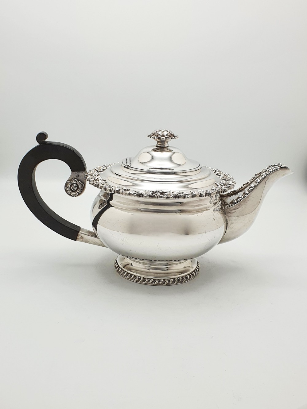 A three-piece silver-plated tea set featuring a teapot with a dark wood handle, a matching sugar bowl with lid, and a cream jug, all decorated with raised grapevine borders. View VII