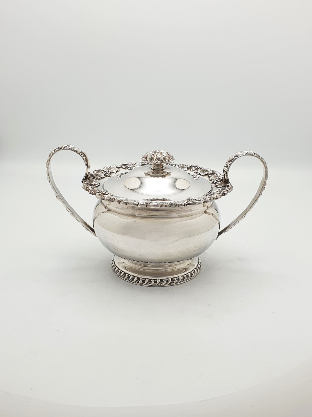 A three-piece silver-plated tea set featuring a teapot with a dark wood handle, a matching sugar bowl with lid, and a cream jug, all decorated with raised grapevine borders. View VIII