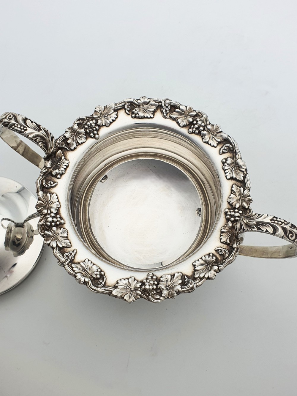 A three-piece silver-plated tea set featuring a teapot with a dark wood handle, a matching sugar bowl with lid, and a cream jug, all decorated with raised grapevine borders. View IX
