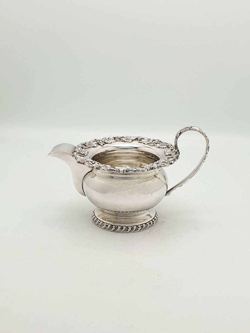 A three-piece silver-plated tea set featuring a teapot with a dark wood handle, a matching sugar bowl with lid, and a cream jug, all decorated with raised grapevine borders. View X