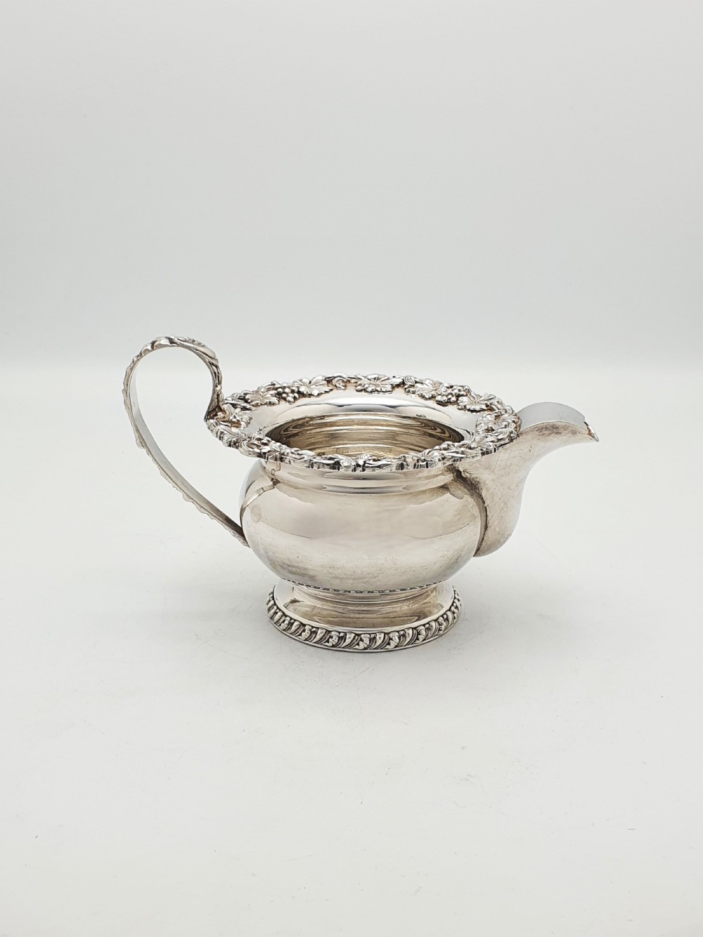 A three-piece silver-plated tea set featuring a teapot with a dark wood handle, a matching sugar bowl with lid, and a cream jug, all decorated with raised grapevine borders. View XI