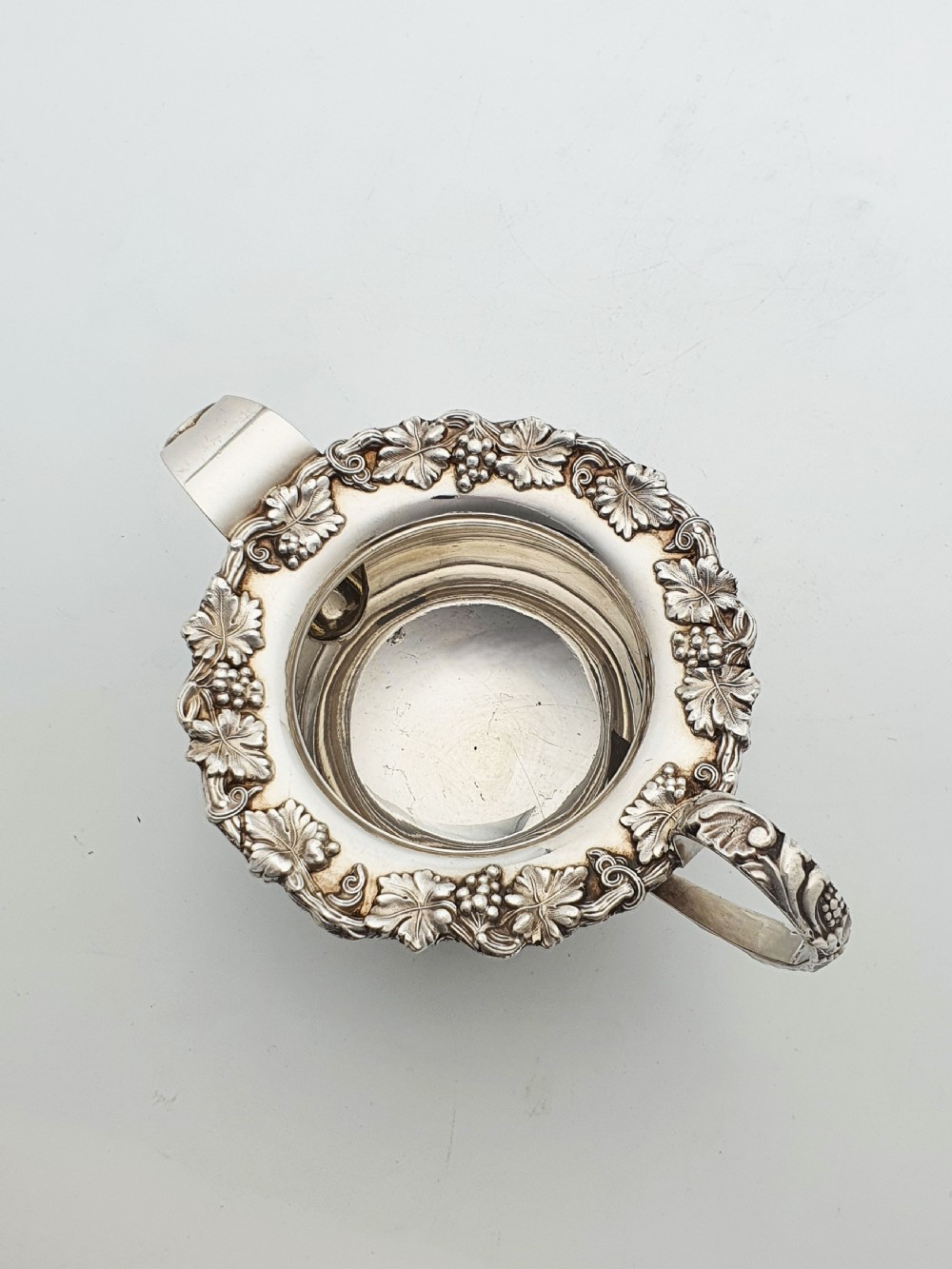 A three-piece silver-plated tea set featuring a teapot with a dark wood handle, a matching sugar bowl with lid, and a cream jug, all decorated with raised grapevine borders. View XII