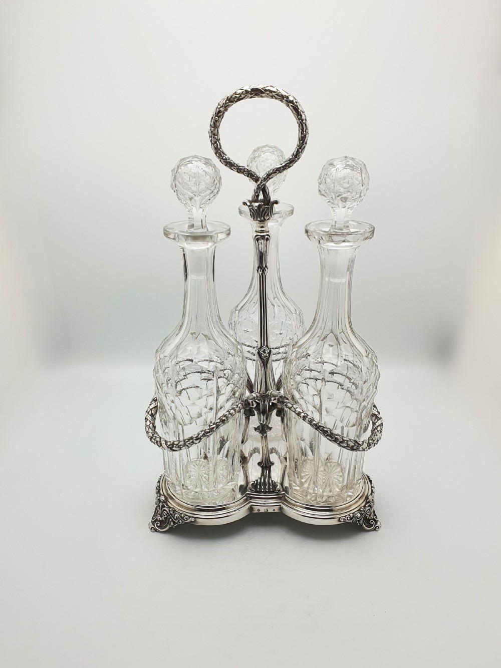 A Victorian silver-plated three-bottle decanter stand holding three clear cut-glass decanters with matching stoppers. View V