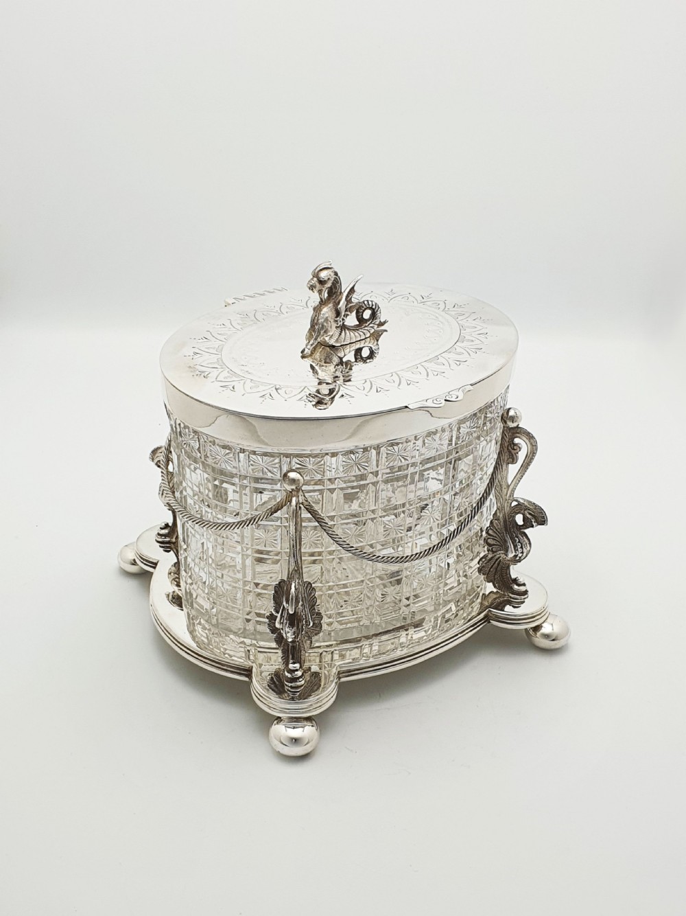 An oval cut crystal biscuit barrel featuring silver-plated mounts, ball feet, twisted rope swags, and an engraved lid topped with a heraldic dragon finial.