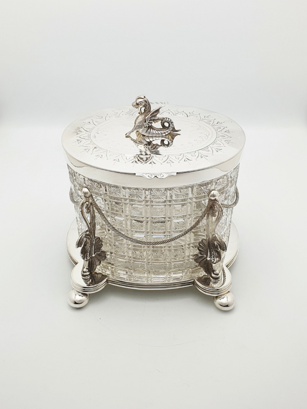 An oval cut crystal biscuit barrel featuring silver-plated mounts, ball feet, twisted rope swags, and an engraved lid topped with a heraldic dragon finial. View II