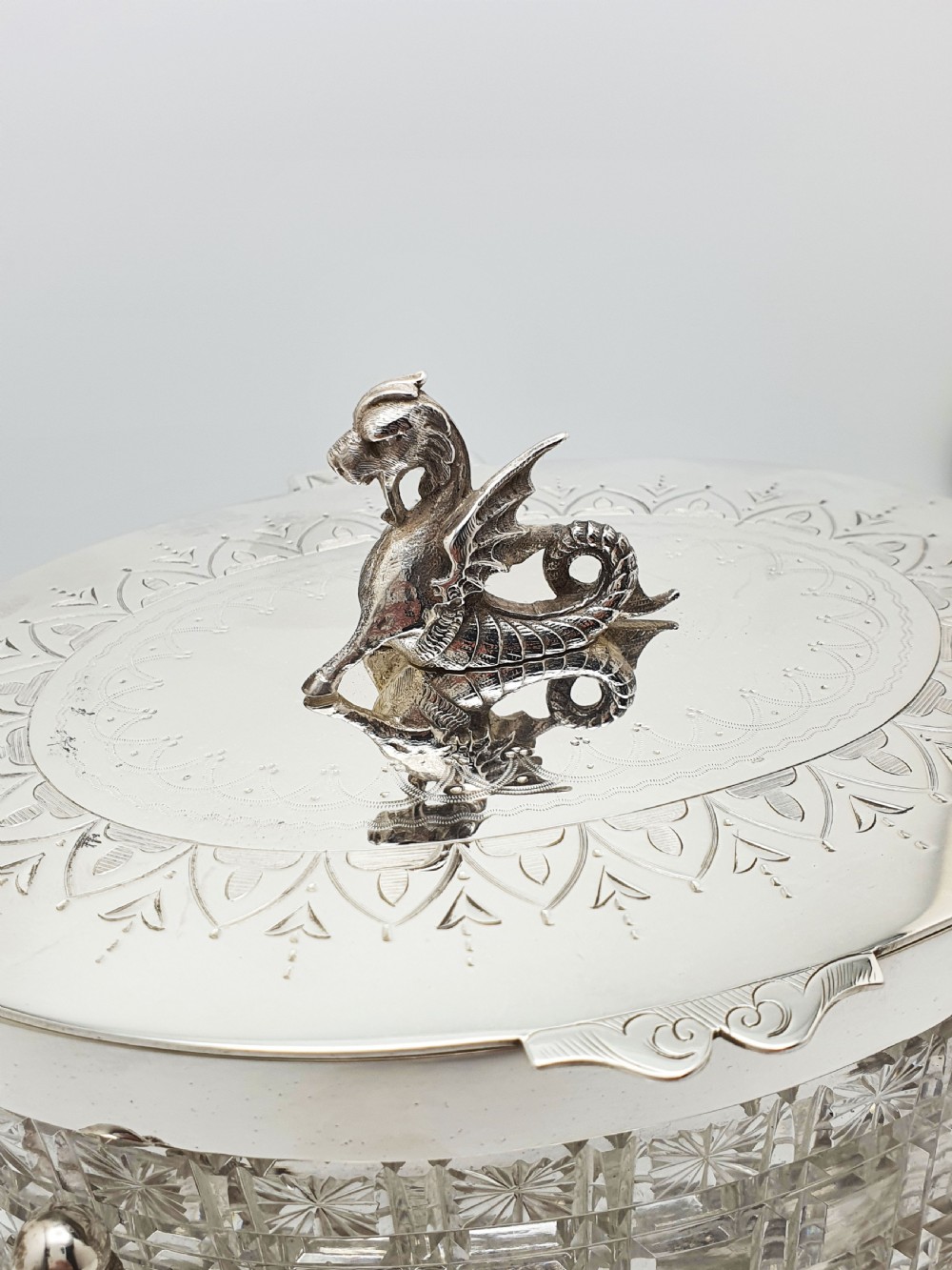 An oval cut crystal biscuit barrel featuring silver-plated mounts, ball feet, twisted rope swags, and an engraved lid topped with a heraldic dragon finial. View V