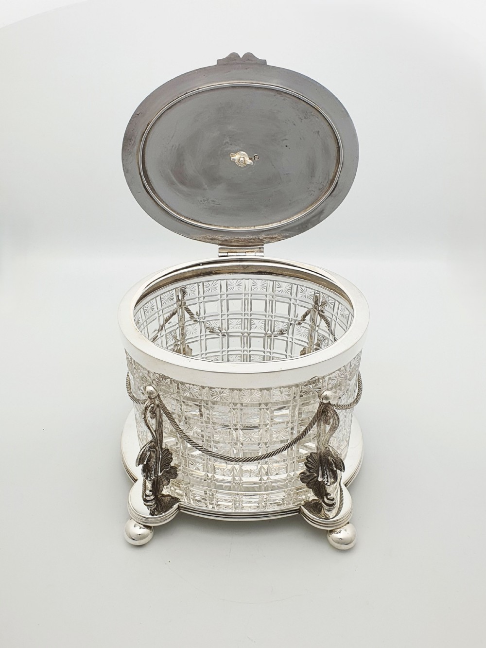 An oval cut crystal biscuit barrel featuring silver-plated mounts, ball feet, twisted rope swags, and an engraved lid topped with a heraldic dragon finial. View VI