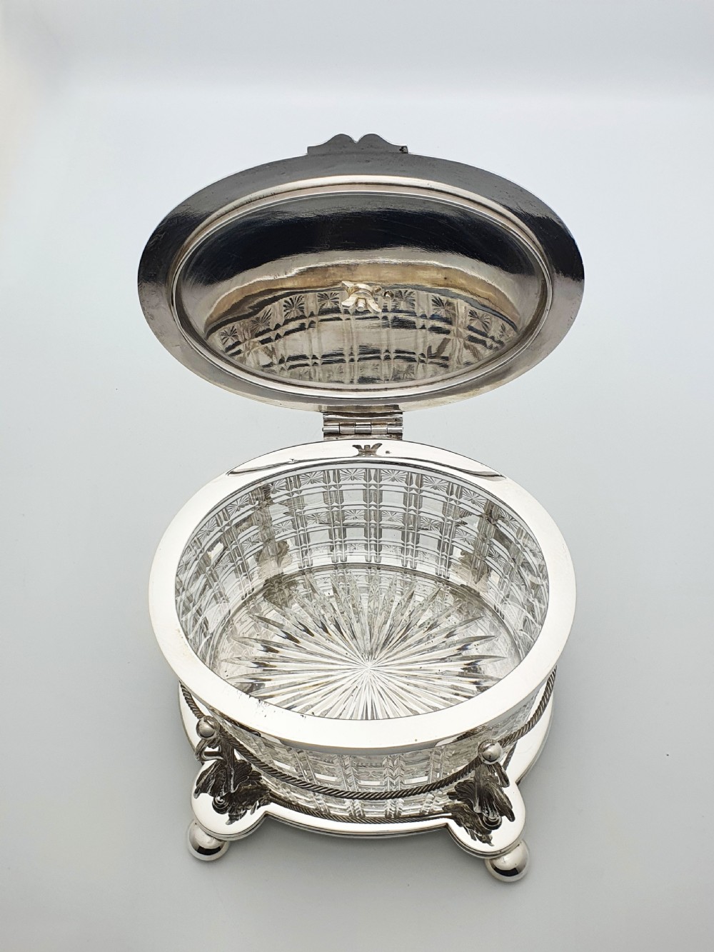 An oval cut crystal biscuit barrel featuring silver-plated mounts, ball feet, twisted rope swags, and an engraved lid topped with a heraldic dragon finial. View VII