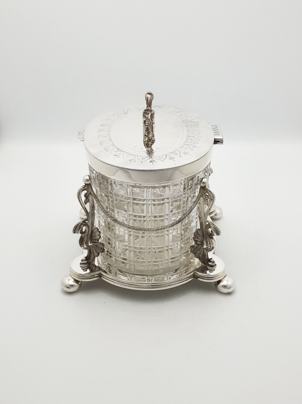 An oval cut crystal biscuit barrel featuring silver-plated mounts, ball feet, twisted rope swags, and an engraved lid topped with a heraldic dragon finial. View VIII