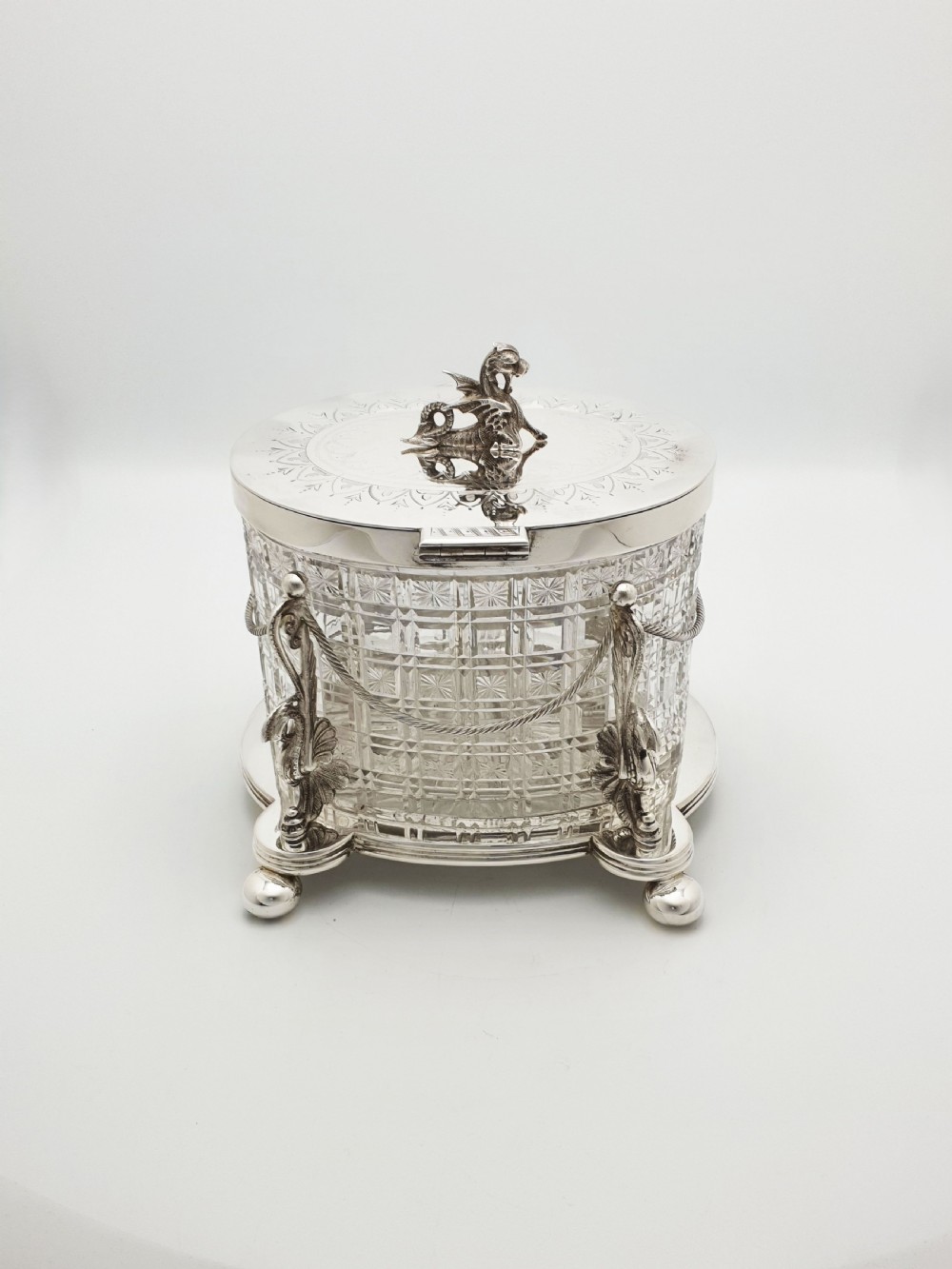 An oval cut crystal biscuit barrel featuring silver-plated mounts, ball feet, twisted rope swags, and an engraved lid topped with a heraldic dragon finial. View IX