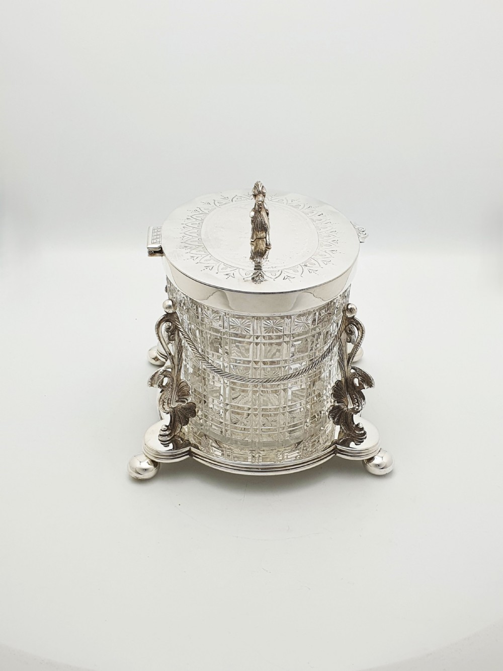 An oval cut crystal biscuit barrel featuring silver-plated mounts, ball feet, twisted rope swags, and an engraved lid topped with a heraldic dragon finial. View X