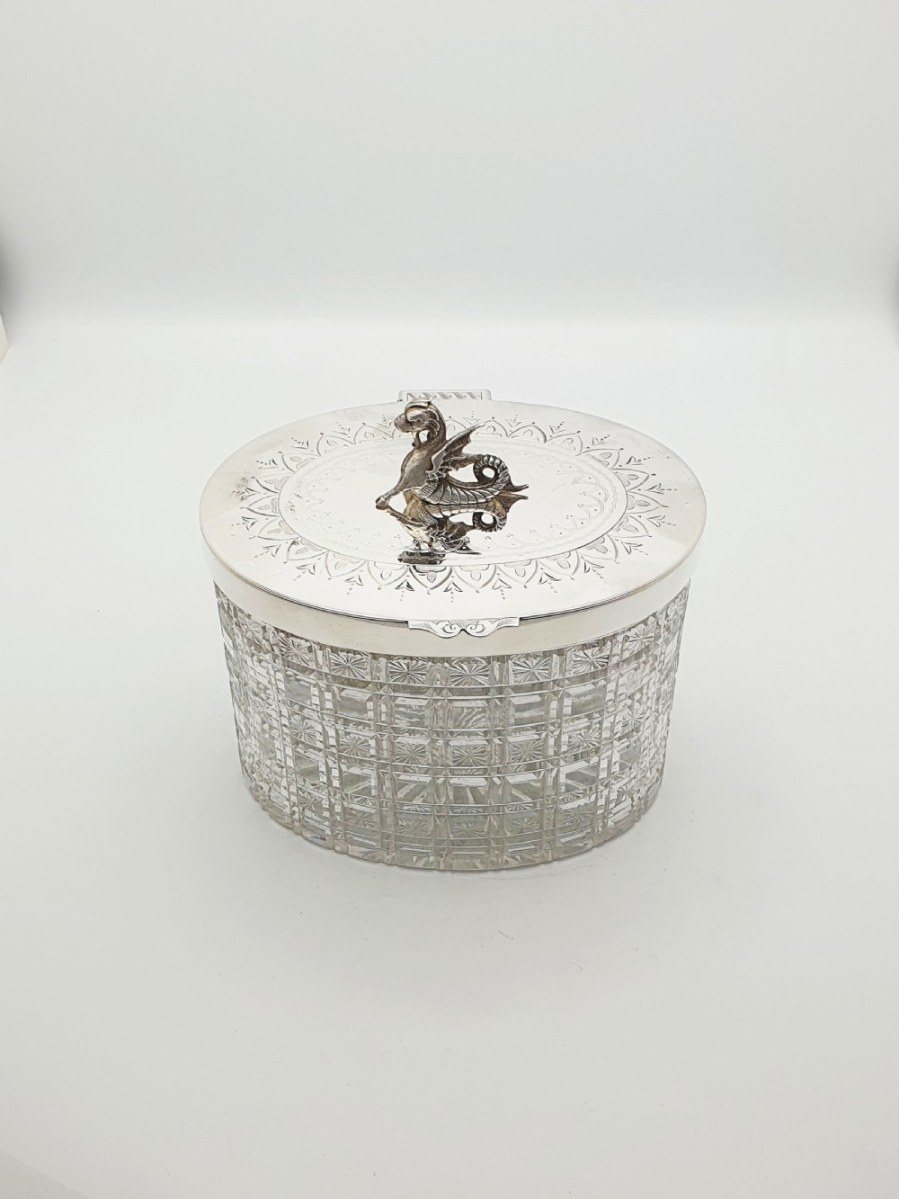 An oval cut crystal biscuit barrel featuring silver-plated mounts, ball feet, twisted rope swags, and an engraved lid topped with a heraldic dragon finial. View XII
